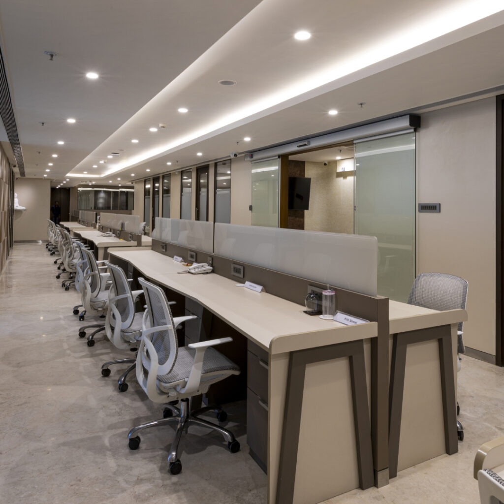 Row of desks with mesh chairs running alongside glass-partitioned cabins, under a cove-lit ceiling